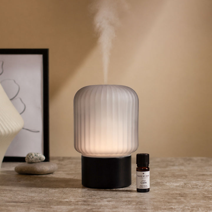 Made By Zen Atmo essential oil diffuser with warm ambient light and aromatic mist on a modern tabletop beside essential oil blend.