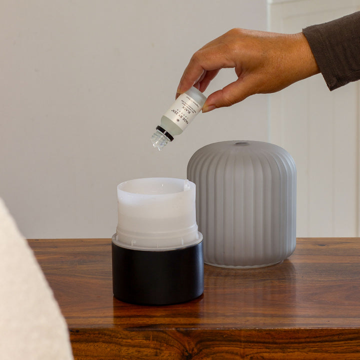 A hand holding an oil bottle above the water tank of the Made By Zen Atmo diffuser on a wooden table. Oil is being added to the water tank. 