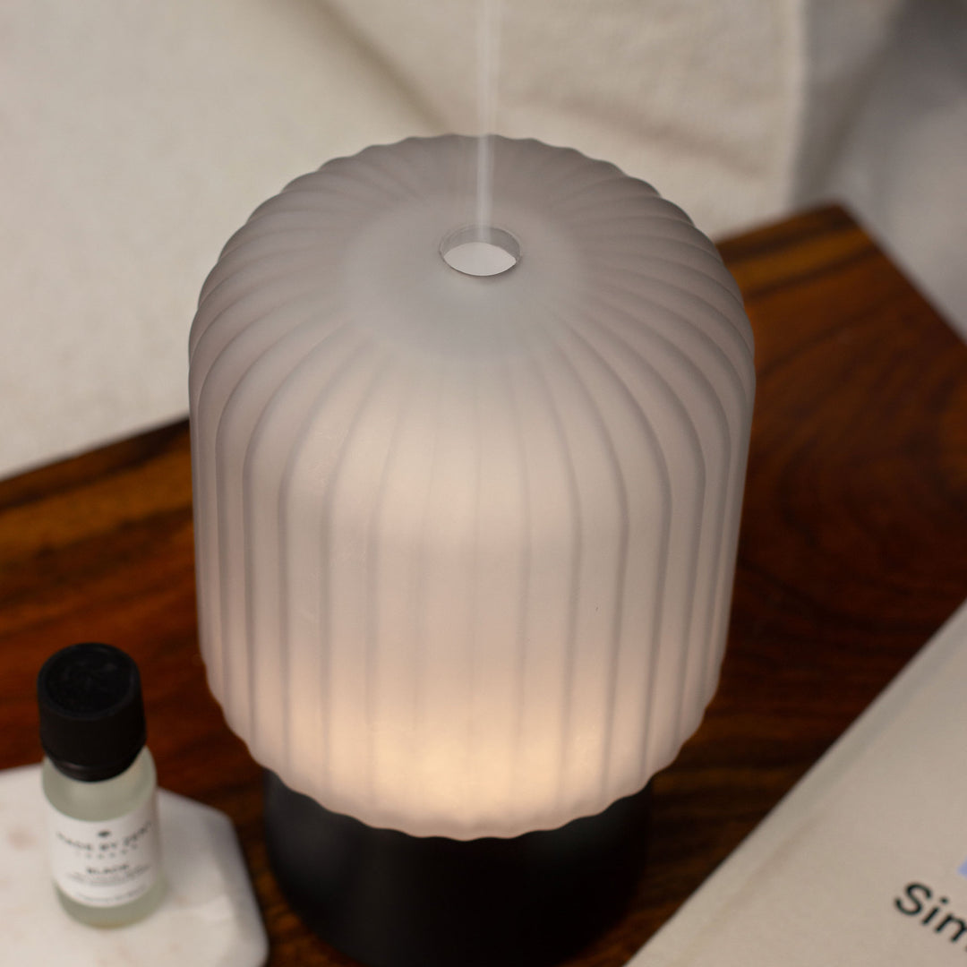 Glass ribbed Made By Zen Atmo diffuser with a black base, emitting a warm glow and a fine mist from the top, shown on a wooden surface next to a small bottle of fragrance oil.