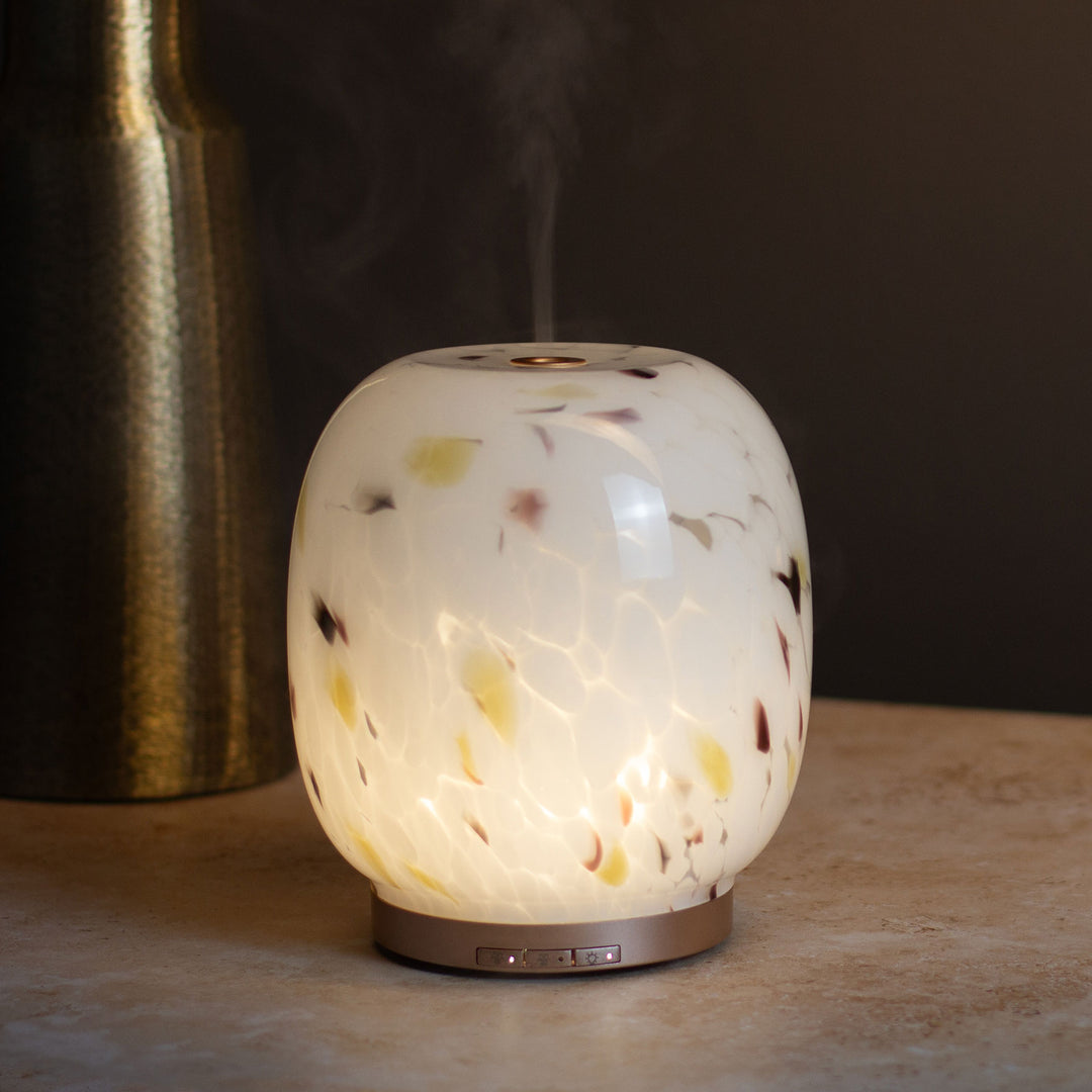 A glowing diffuser lamp with a white confetti glass finish on a stone surface with a gold vase against a dark background.