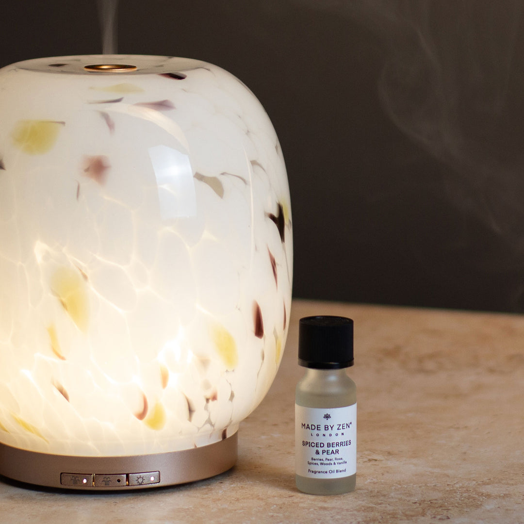 A glowing diffuser lamp with a white confetti glass finish next to a Made by Zen fragrance oil on a stone surface against a dark background.