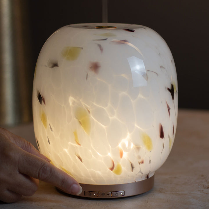 A glowing glass diffuser with a hand touching its base, featuring a confetti glass finish with brown and amber flecks emitting a warm light.