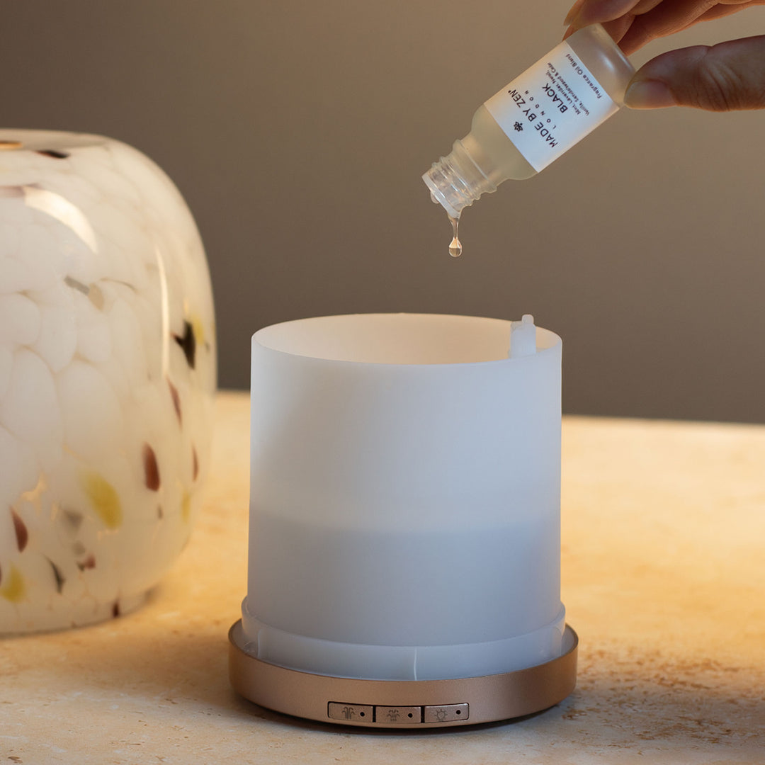 Hand pouring liquid from a Made by Zen fragrance oil into the water tank of an aroma diffuser. With the confetti glass cover in the background.