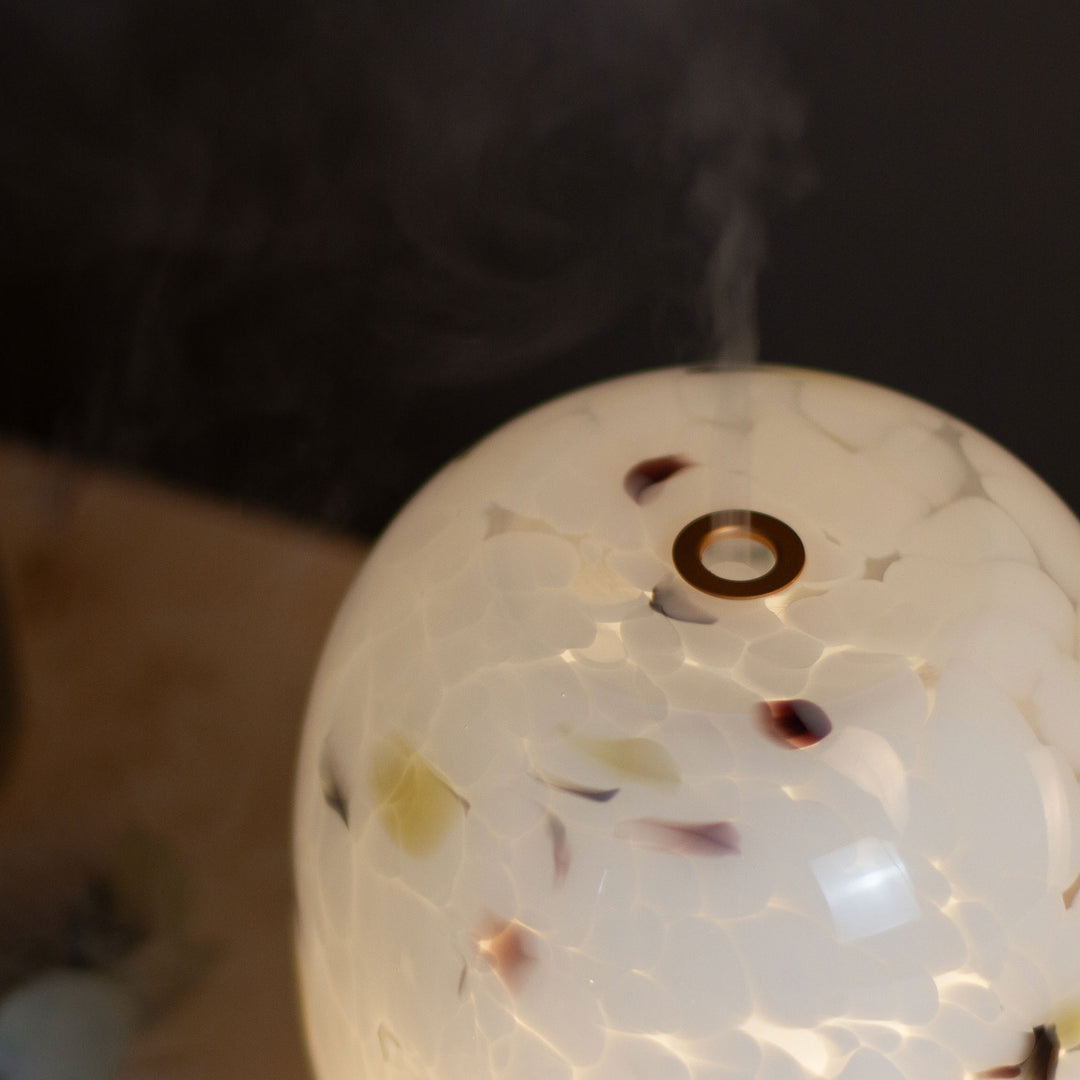 A close-up of a confetti glass aroma diffuser showing themist rising from the opening against a dark background.