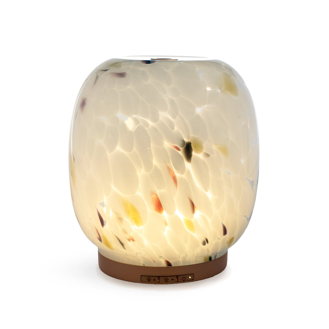 A glowing aroma diffuser with a rounded, glossy glass with confetti effect, on a metallic gold base against a white background.